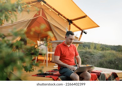 Happy man freelancer programmer using a laptop on a cozy glamping tent in a summer day. Luxury camping tent for outdoor holiday and vacation. Lifestyle concept - Powered by Shutterstock