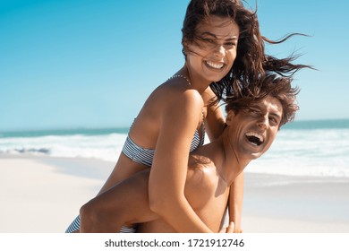 Happy man giving piggyback ride to his woman and laughing at tropical beach. Smiling guy in love carrying on back her girlfriend and having fun. Joyful couple enjoying summer at sea with copy space.