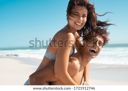 Happy man giving piggyback ride to his woman and laughing at tropical beach. Smiling guy in love carrying on back her girlfriend and having fun. Joyful couple enjoying summer at sea with copy space.