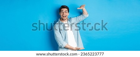 Happy man in glasses showing big size object, shaping large box, standing over blue background and smiling.