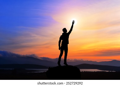 happy man with hand up on sunset background, man on top of the mountain reaches for the sun, silhouette man holding sun on the hill