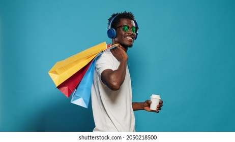 Happy man with handbags dancing after shopping spree. Cheerful person with sunglasses listening to music on headphones while holding bags with clothing for sale purchase on black friday - Powered by Shutterstock