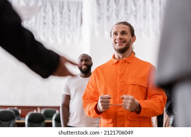 Happy Man In Handcuffs And Jail Uniform Near Blurred Jurors In Courtroom