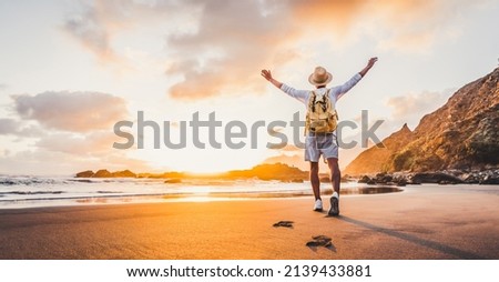 Happy man with hands up enjoying wellbeing and freedom at the beach - Male with backpack traveling in the nature with sunrise view - Healthy lifestyle, happiness and travel concept