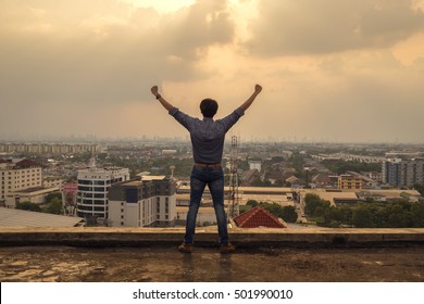 Happy man with hands up on sunset. Open arms, standing on a pier barefoot. Enjoying sun. Gazing sun. top tower looking city