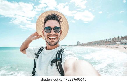 Happy man with hat and sunglasses taking selfie picture with smartphone at the beach - Cheerful traveler having fun outside - Handsome guy smiling at camera - Powered by Shutterstock