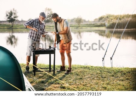 Happy man and his senior father preparing meat on barbecue grill while camping by the river in nature. Copy space.
