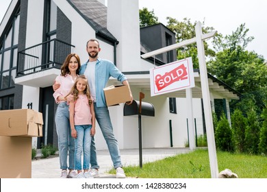 Happy Man Holding Box And Standing With Wife And Daughter Near House And Board With Sold Letters 