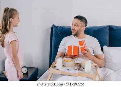 Happy Man Holding Fathers Day Gift Boxes While Having Breakfast In Bed Near Adorable Daughter