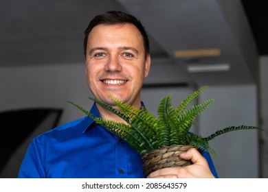 Happy Man Holding Fern House Plant. Successful Millennial Man, Dressed In Blue Shirt Holding Flower Pot With Green Fern Plant House And Looks At Camera