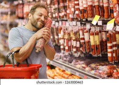 Happy man holding in hands near face salami and smiling. Bearded customer standing with closed eyes in meat section. Handsome man choosing delicious meat of whole assortment. - Powered by Shutterstock