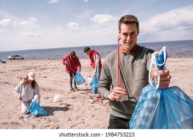 Happy Man Holding Trash Bag Near Group Of Blurred Volunteers Picking Up Rubbish On Sand 