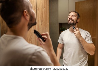 A happy man is holding trimmer and preparing to shave his beard in bathroom. - Powered by Shutterstock