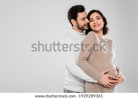 Happy man hugging cheerful pregnant woman isolated on grey 
