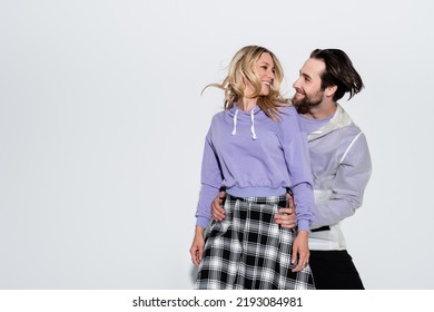 Happy Man Hugging Cheerful Woman In Purple Sweatshirt And Skirt While Looking At Each Other On Grey