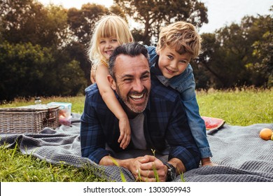 Happy Man With Kids On A Picnic Lying Down In Park Beside A Picnic Basket. Children Lying On The Back Of Their Father.