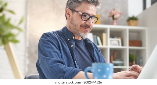 Happy Man With Laptop Computer At Home. Businessman Working At Desk In Home Office. Portrait Of Mature Age, Middle Age, Mid Adult Man, Bearded, Glasses, Smiling, Authentic Look.