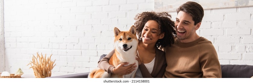 Happy man looking at smiling african american girlfriend with shiba inu at home, banner - Powered by Shutterstock