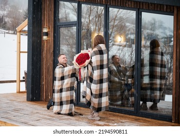 Happy Man In Love Making Marriage Proposal To Girlfriend Under Winter Snow While Woman Holding Bouquet Of Flowers. Couple Sharing Romantic Moment Outside Scandinavian House Barnhouse.