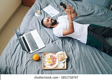 Happy man lying on bed with tray with breakfast, laptop, coffee cup and report around him and texting friends - Powered by Shutterstock