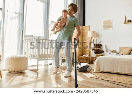 happy man multitasking housework and childcare, father vacuuming hardwood floor with infant in arms