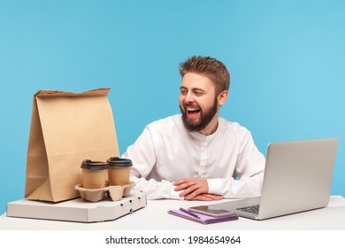 Happy man office worker looking at pizza box, cups of coffee and paper bag with purchase with smile, enjoying fast food delivery. Indoor studio shot isolated on blue background - Powered by Shutterstock