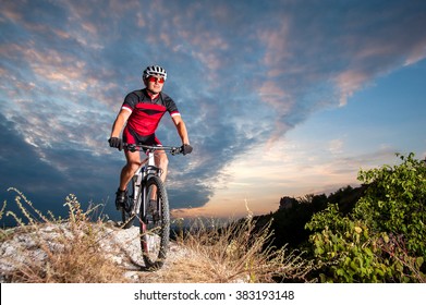 Happy Man On A Mountain Bike Races Downhill In The Nature Against Blue Cloudy Evening Sky. Cyclist Is Wearing Red Sportswear Helmet Gloves And Red Glasses. Cross Country Biking.