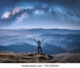 Happy man on the mountain peak and arched Milky Way over mountains in low clouds at night. Landscape with blue sky with stars, Milky Way Arch, guy, hills in fog. Space and galaxy. Sky with stars