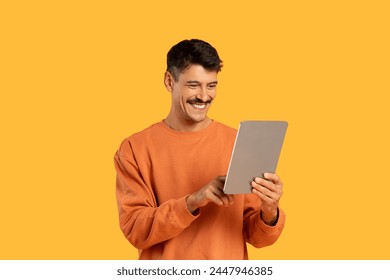 Happy man in an orange sweater looking at a tablet screen with a joyful expression on a yellow background - Powered by Shutterstock