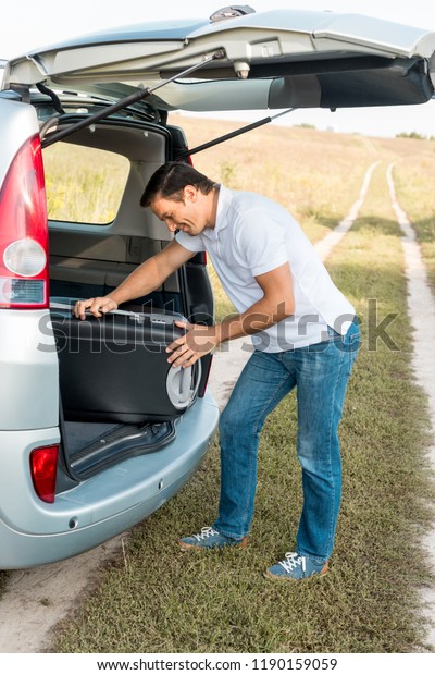 happy man packing suitcase into luggage boot of car\
in field