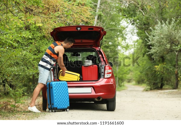 Happy man packing suitcases into car trunk near\
forest. Space for text