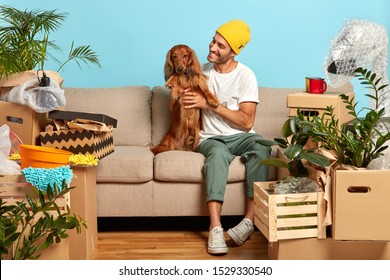Happy man pets pedigree dog, pose on sofa, move to new house, lot of packing cartons around, rejoice buying modern flat, rest after moving in. Homeowner with pet spend first day at new dwelling - Shutterstock ID 1529330540