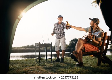 Happy man playing acoustic guitar and toasting with his father who is grilling meat during their camping day. - Powered by Shutterstock