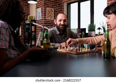 Happy man playing chess with multiethnic friends at home in living room. Diverse people sitting at table and having fun while playing boardgames together and enjoying snacks and beverages.