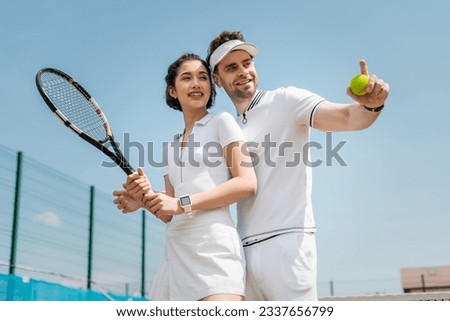 happy man pointing away near girlfriend on tennis court, holding rackets , sport and romance