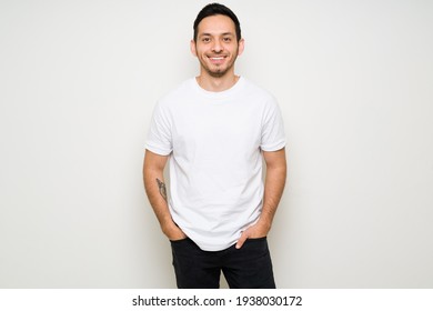 Happy Man Posing With The Hands In His Pockets And Smiling. Latin Man Wearing A Mock Up T-shirt Design While Standing In Front Of A White Background