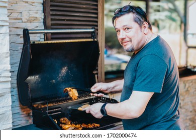 Happy Man Preparing Food On Barbecue. An Older Guy Grilling Meat On Grill For Family And Friends. Sunny Day For Party Bbq. Senior Rests And Doing Meal Grilling In Beautiful Weather.