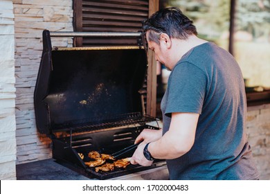 Happy Man Preparing Food On Barbecue. An Older Guy Grilling Meat On Grill For Family And Friends. Sunny Day For Party Bbq. Senior Rests And Doing Meal Grilling In Beautiful Weather. 