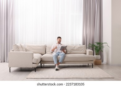 Happy man reading book on sofa near window with beautiful curtains in living room - Powered by Shutterstock