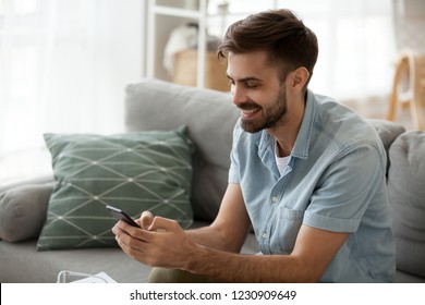 Happy man relax on couch at home chatting with friends on smartphone, excited millennial male writing message on cell to girlfriend, smiling guy using mobile phone texting or playing app games