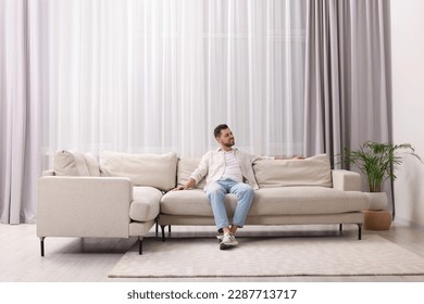 Happy man resting on sofa near window with beautiful curtains in living room - Powered by Shutterstock