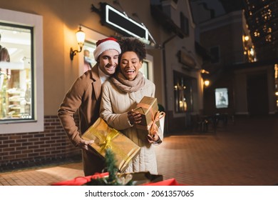 happy man in santa hat and african american girlfriend looking at blurred shopping bags while holding christmas gift boxes - Powered by Shutterstock