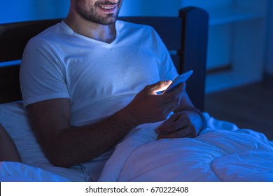 The Happy Man Sit With A Phone In The Bed. Night Time