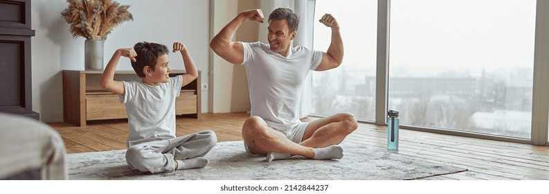 Happy man is sitting with boy on mat and showing biceps after doing workout together - Powered by Shutterstock