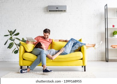 happy man with smiling girlfriend relaxing on yellow sofa under air conditioner at home