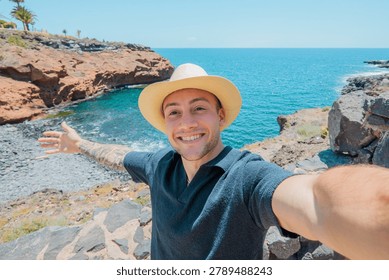 Happy man smiling while taking a selfie standing on a rocky coast with an arm outstretched, enjoying a summer vacation by the blue ocean and cliffs under a clear sky