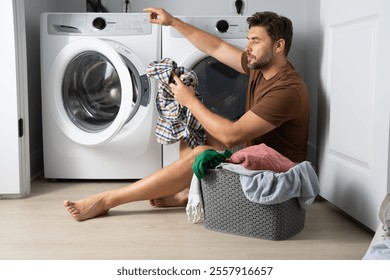 Happy man sorting clothes near washing machine in laundry room. man sits on the floor of a house near a washing machine with dirty clothes. a man does his laundry at home. a man holding dirty clothes - Powered by Shutterstock