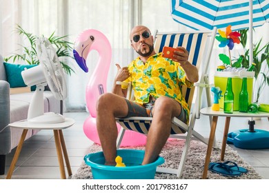 Glücklicher Mann, der die Sommerferien zu Hause verbringt und so tut, als wäre er am Strand, sitzt er auf einem Liegestuhl und macht sich mit seinem Smartphone selbstsüchtig
