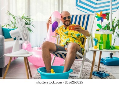 Happy Man Spending Summer Vacations At Home And Pretending He Is On A Beach, He Is Sitting On A Deckchair And Taking Selfies With His Smartphone