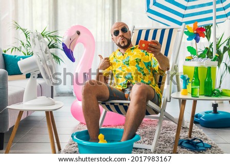 Happy man spending summer vacations at home and pretending he is on a beach, he is sitting on a deckchair and taking selfies with his smartphone